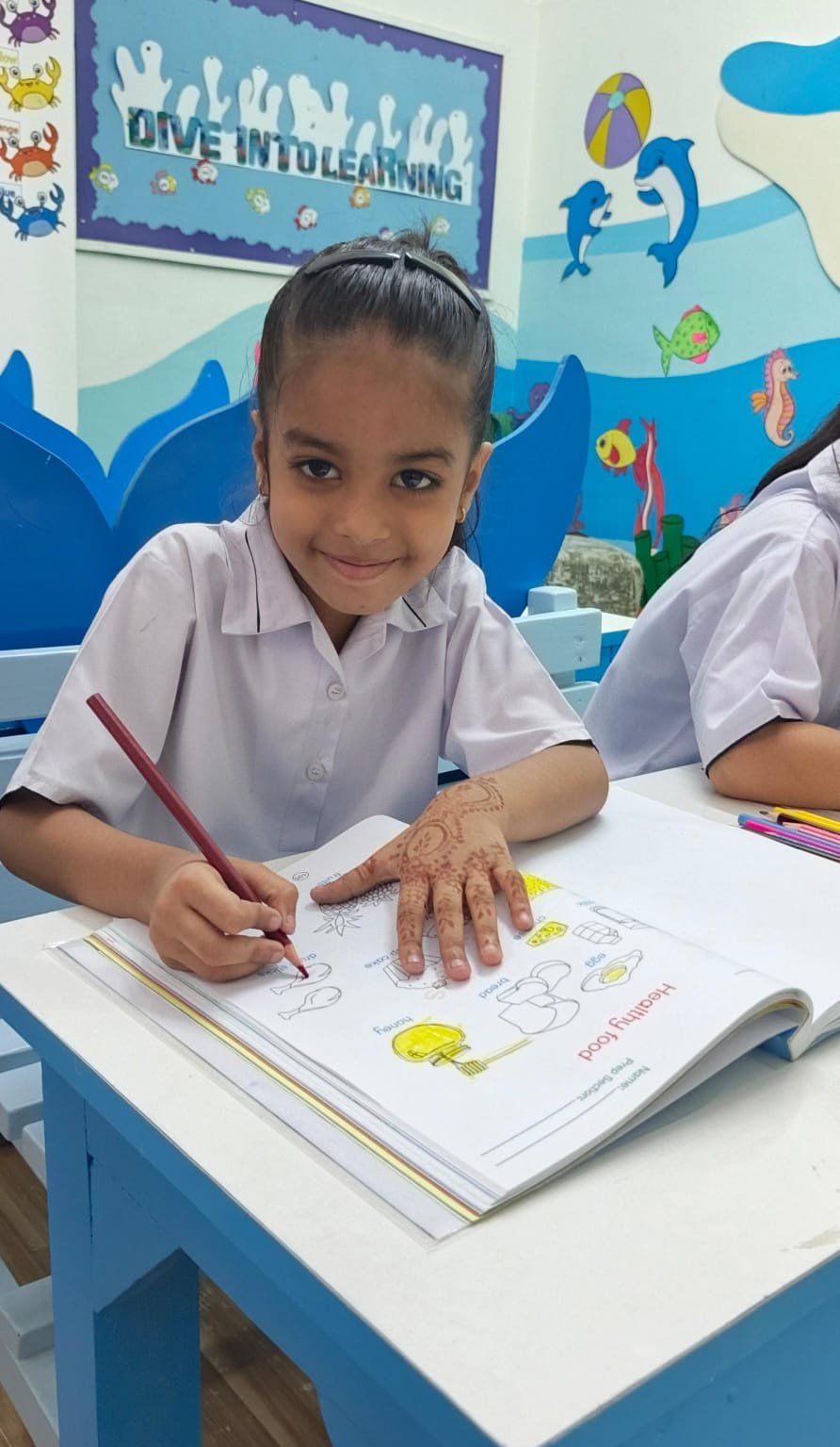 A Warsis student smiling while colouring a healthy-food worksheet