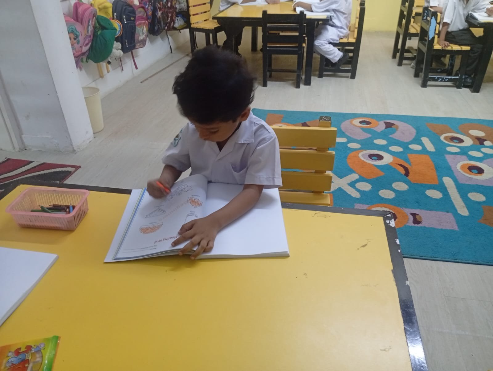 A Warsis student colouring at a classroom table with classmates behind him